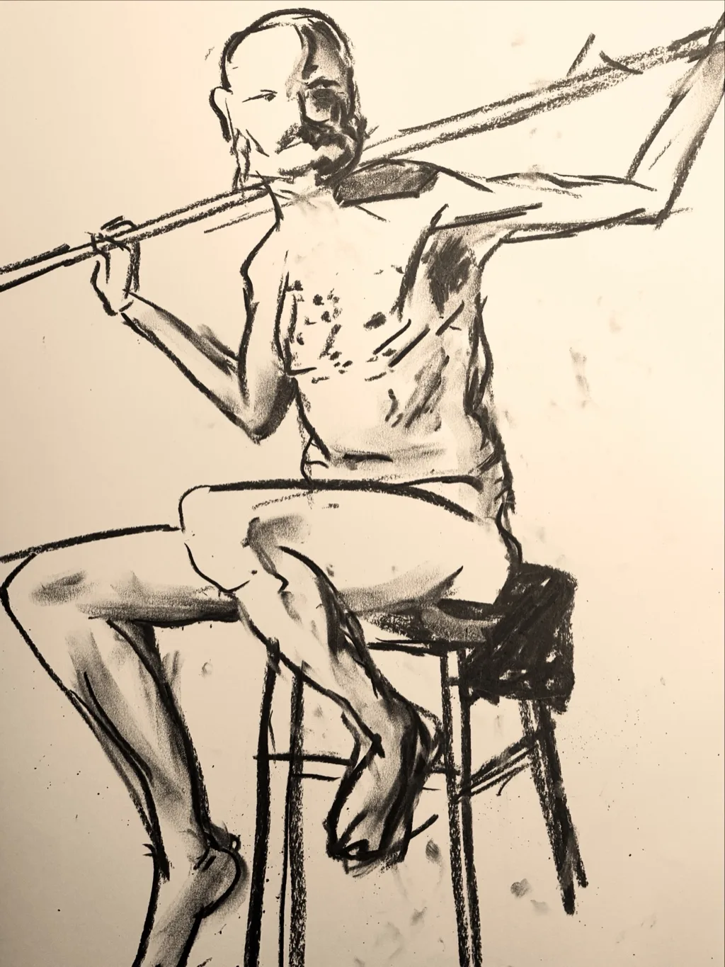 Charcoal figure drawing of a seated nude model on a stool holding a staff