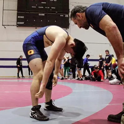 Wrestler in a Queen's singlet bending over with a coach on the mat